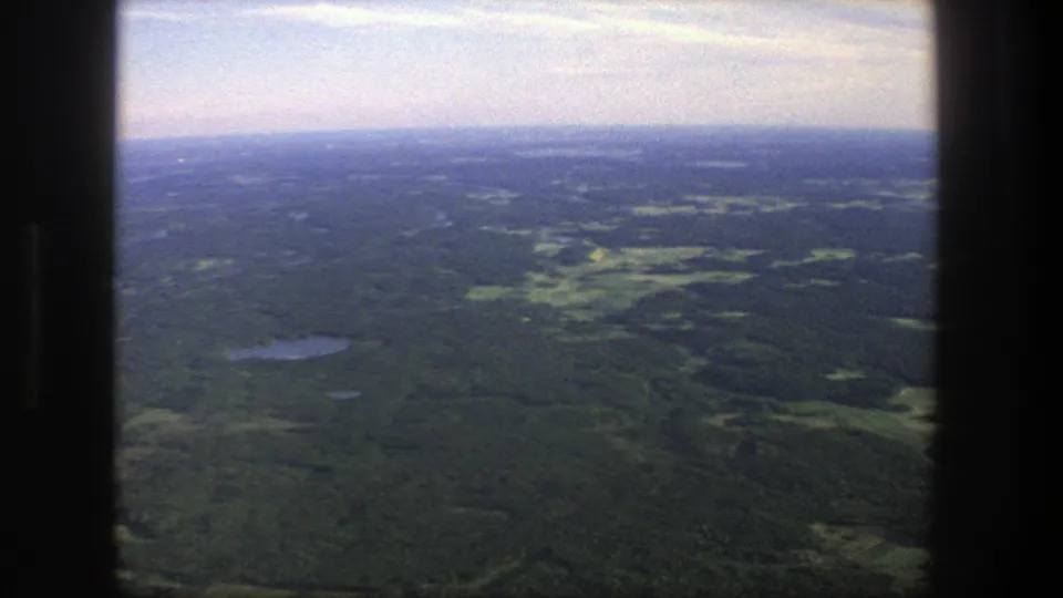 1975 Denmark home-movie frame showing a broad aerial landscape of forest, small lakes, and light fields beneath a hazy sky.