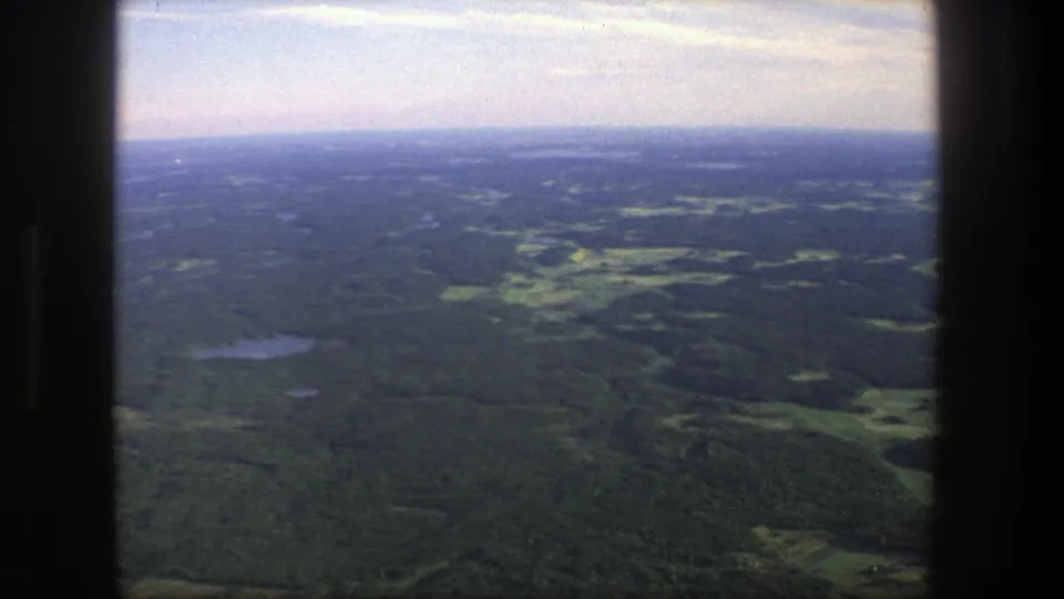 1975 Denmark 8mm frame from an aircraft window, with wooded countryside, small lakes, field clearings, and a soft hazy horizon.