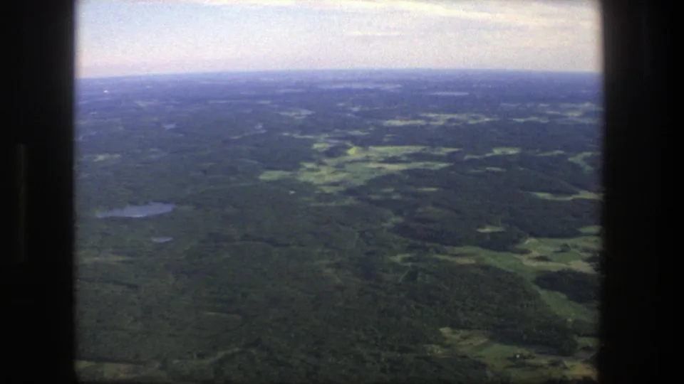 1975 Denmark aerial home-movie frame showing green forest, scattered small lakes, open fields, and a hazy blue horizon.