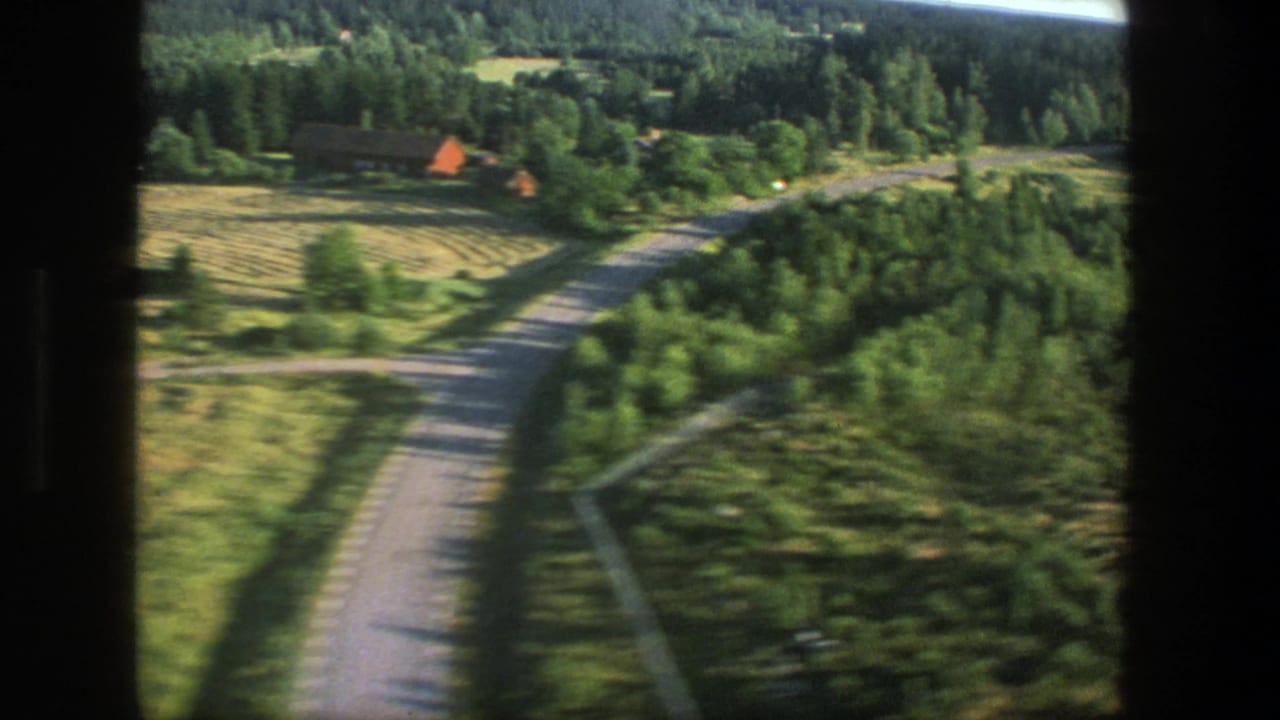 1975 Denmark 8mm passenger-window view over green countryside, a winding road, forest edges, and a distant red-roofed structure.