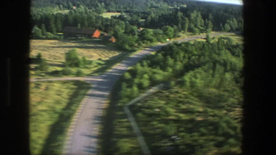 1975 Denmark 8mm passenger-window view over green countryside, a winding road, forest edges, and a distant red-roofed structure.