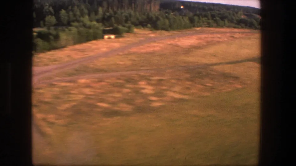 Motion-blurred 1975 Denmark 8mm frame from a descending aircraft, showing open rural ground and forest edge below.