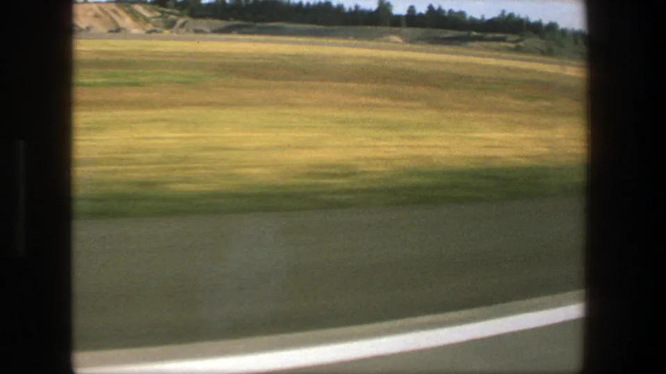 1975 Denmark 8mm frame showing blurred roadside pavement, green-yellow fields, and a dark tree line from a passenger perspective.