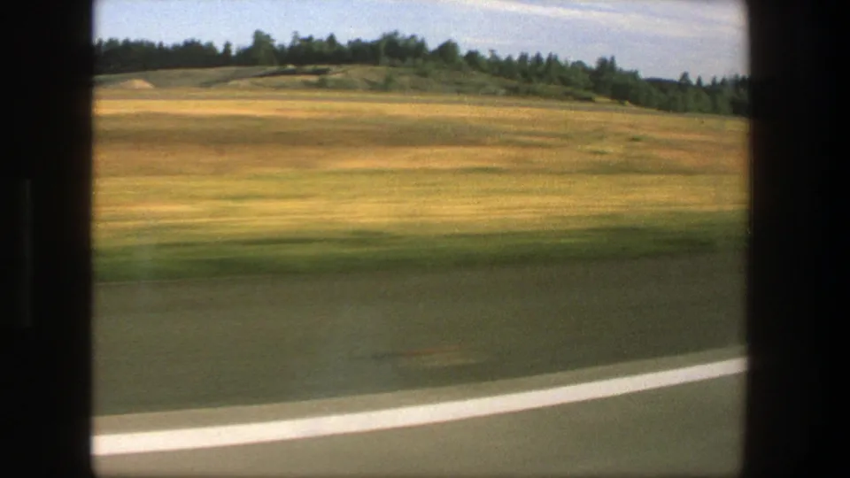 1975 Denmark 8mm frame showing a white road edge, blurred fields, low hills, and trees from a moving vehicle.