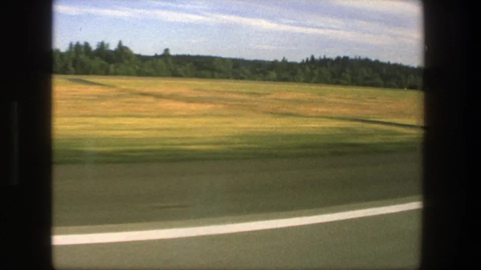 1975 Denmark countryside view with a curving road edge, green and gold fields, wooded horizon, and film-gate framing.