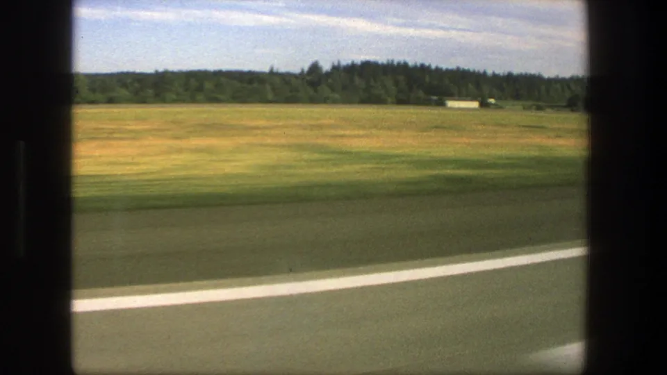 1975 Denmark rural travel frame with blurred road edge, broad fields, a distant treeline, and a small pale structure near the horizon.