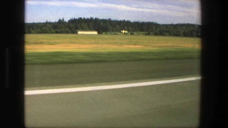 1975 Denmark countryside frame showing a small white building across open fields, with road edge blur and dark window framing.