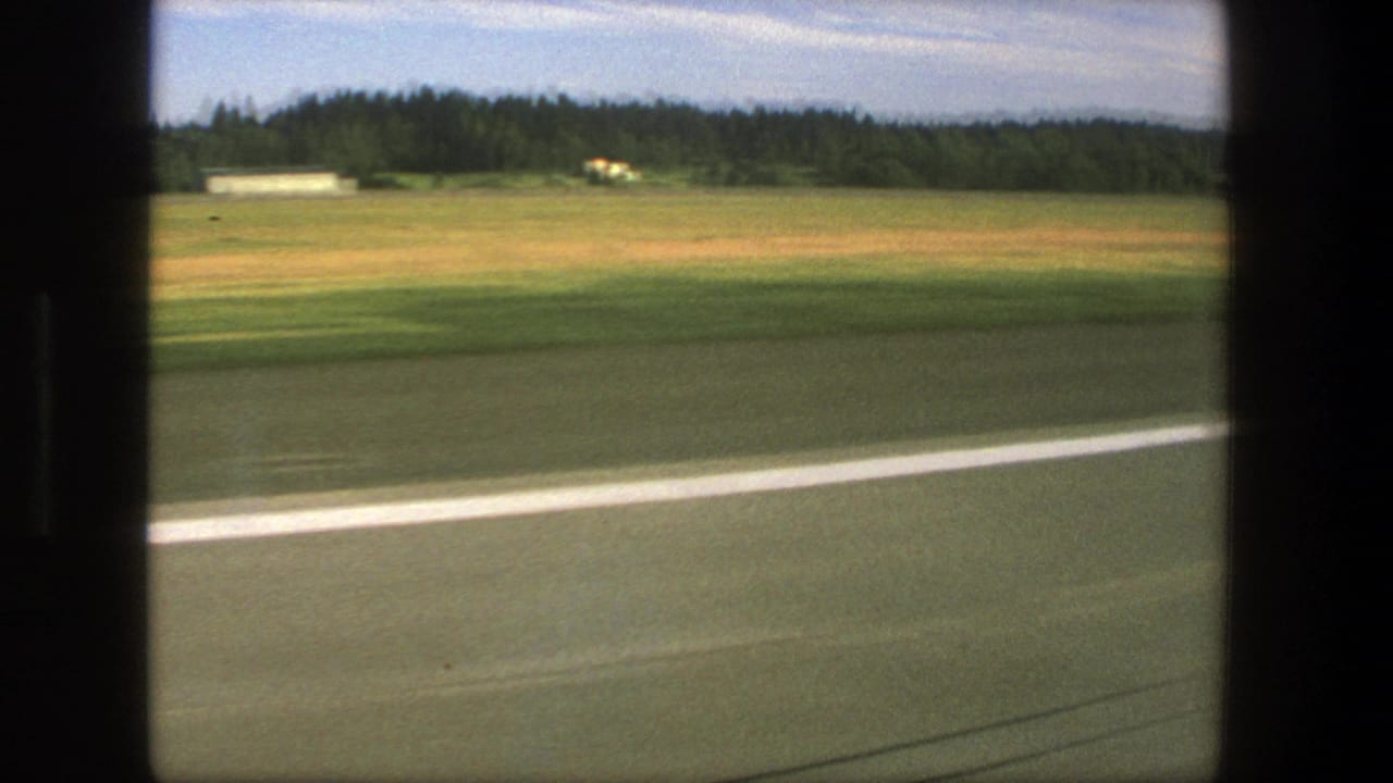 1975 Denmark 8mm road view from a moving vehicle, showing pavement, a white line, open field, trees, and a small distant building.