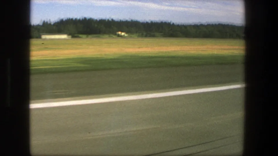 1975 Denmark 8mm road view from a moving vehicle, showing pavement, a white line, open field, trees, and a small distant building.