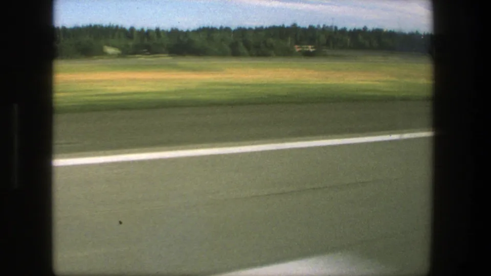 1975 Denmark roadside scene with a white road line, open field, wooded background, and small distant structures filmed in motion.
