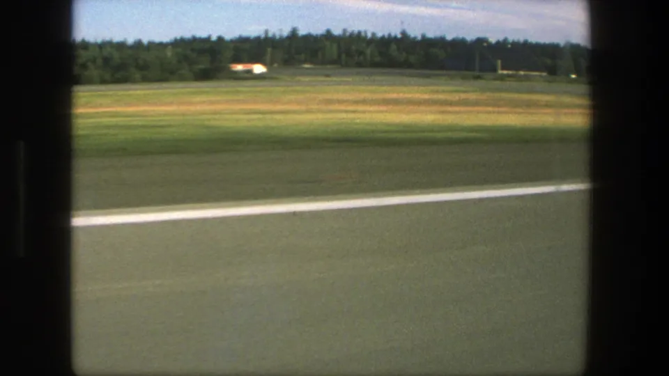 Moving 8mm view of a Danish roadside in 1975, showing open grass, pavement, a white line, distant buildings, and trees.