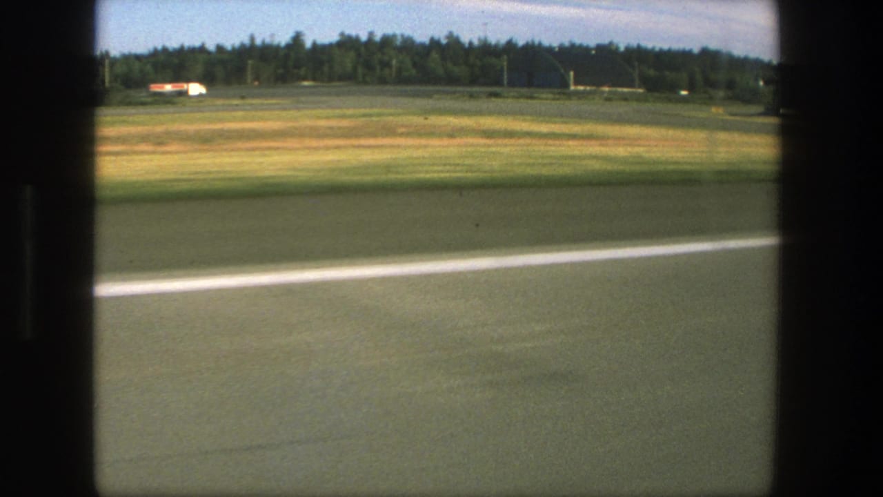 1975 Denmark home-movie view from a plane window showing runway pavement, grass, treeline, and distant airfield structures.