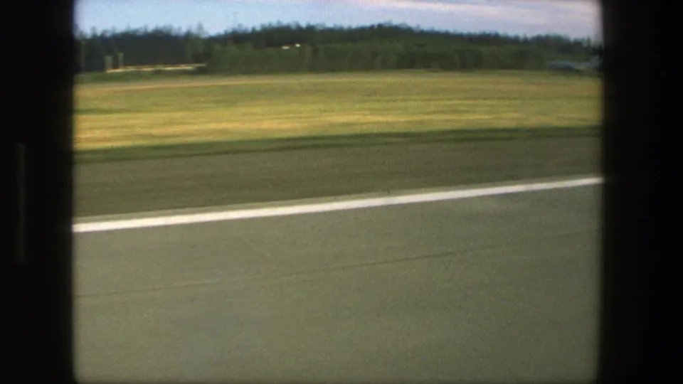 1975 Denmark home-movie frame showing runway markings, open grass, treeline, and airport buildings from a moving aircraft.