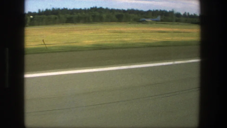 1975 Denmark passenger-window view with runway pavement, grassy field, treeline, and a possible distant aircraft near the airport edge.