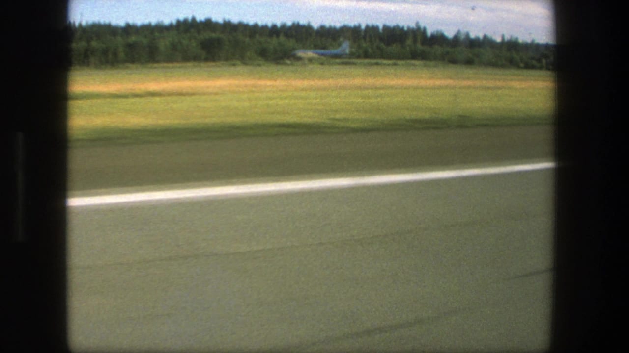 1975 Denmark airport view from a moving vehicle, showing runway pavement, a grassy field, treeline, and a distant parked airplane.