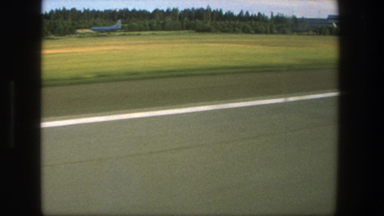 Runway pavement and white stripe pass in the foreground with grass, trees, and a small aircraft in the distance.