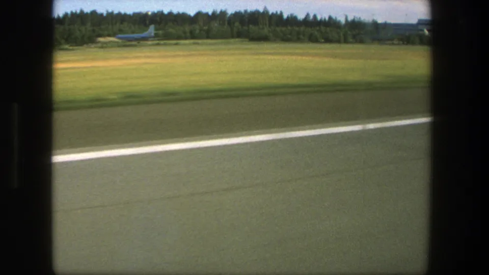 Moving 8mm airport view in Denmark, with runway edge, open grass, forested background, and a distant airplane near the treeline.