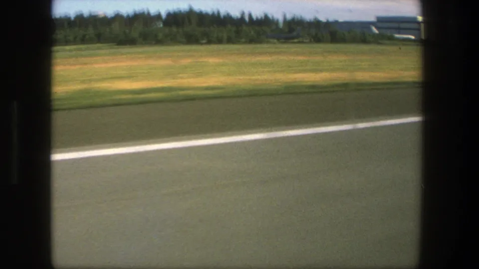 1975 Danish airport ground view from a moving vehicle, showing runway pavement, grass, treeline, distant aircraft, and low airport structures.
