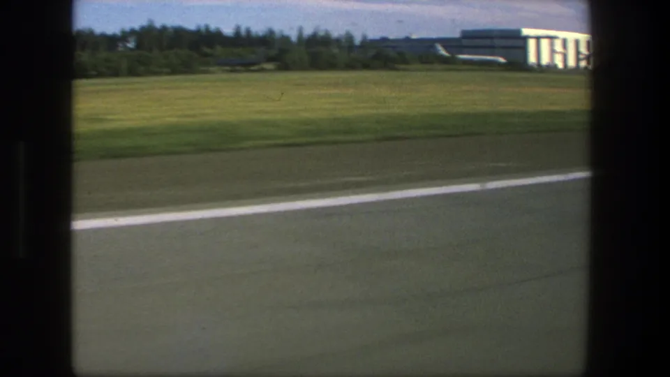 Moving airport view in 1975 Denmark, with runway pavement, grassy field, trees, low buildings, and partial distant aircraft visible.
