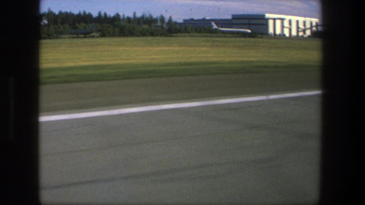 1975 Denmark airport view from inside an aircraft, showing runway pavement, grass, and a hangar-like building beyond the window frame.