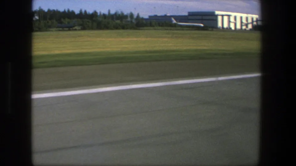 1975 Denmark airport view from inside an aircraft, showing runway pavement, grass, and a hangar-like building beyond the window frame.