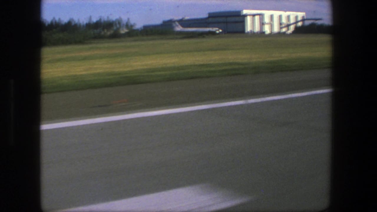 Airport runway and grassy field pass by outside an aircraft window, with a large building beyond the field.