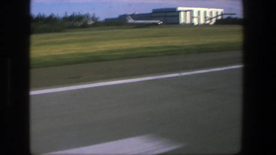 A handheld 8mm view from an aircraft window in Denmark, 1975, with runway markings, grass, and a distant airport building.