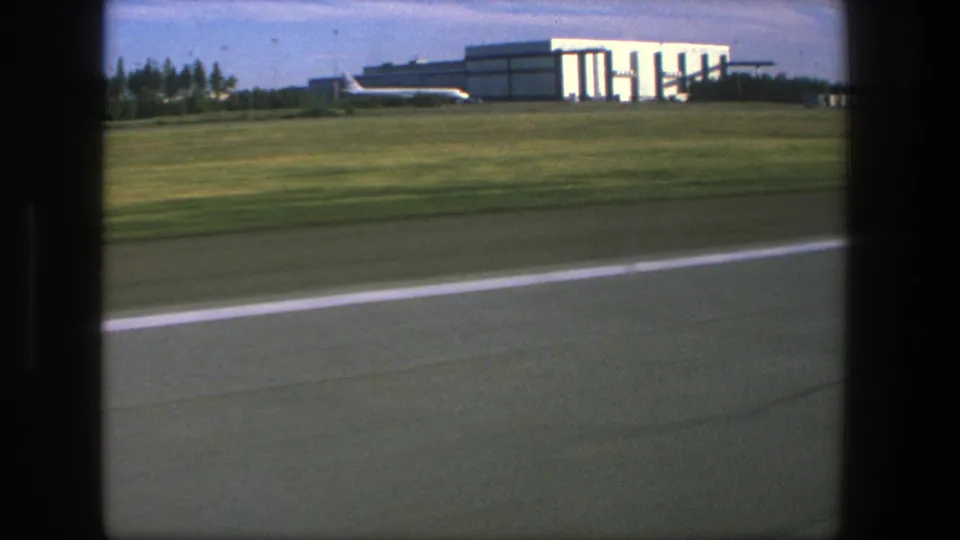 1975 Denmark airport tarmac view through an aircraft window, with a broad grass verge and hangar-like building under blue sky.