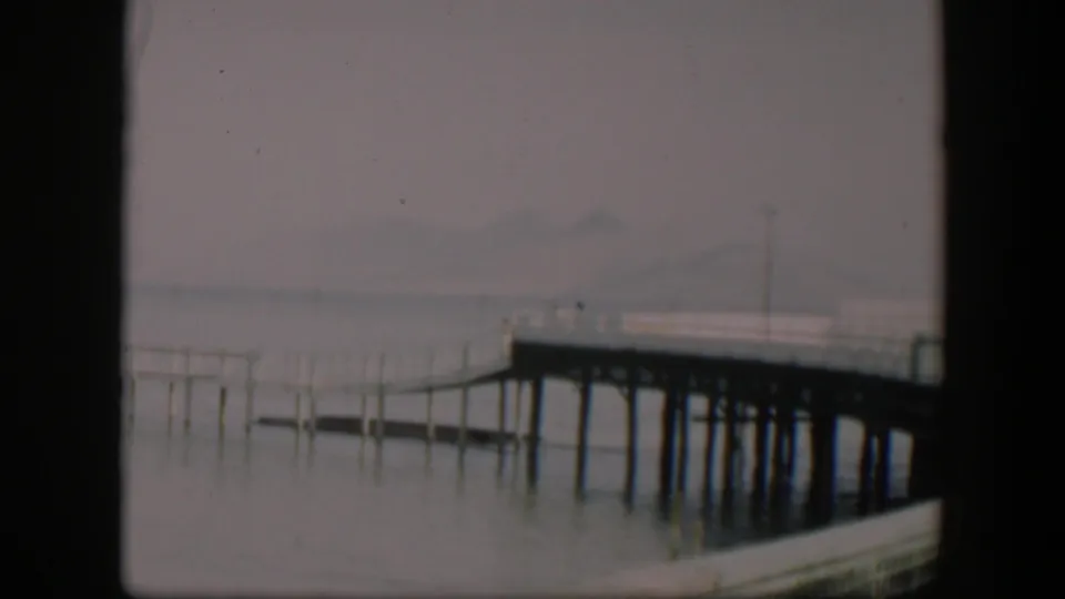 Vintage 8mm frame of the same hazy waterside dock, with pilings and faint mountains softened by exposure and age.
