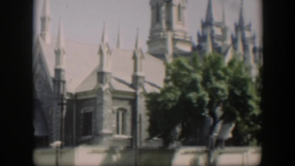 1951 Wyoming home-movie frame showing the side of a stone church, pointed spires, leafy trees, and heavy dark frame borders.
