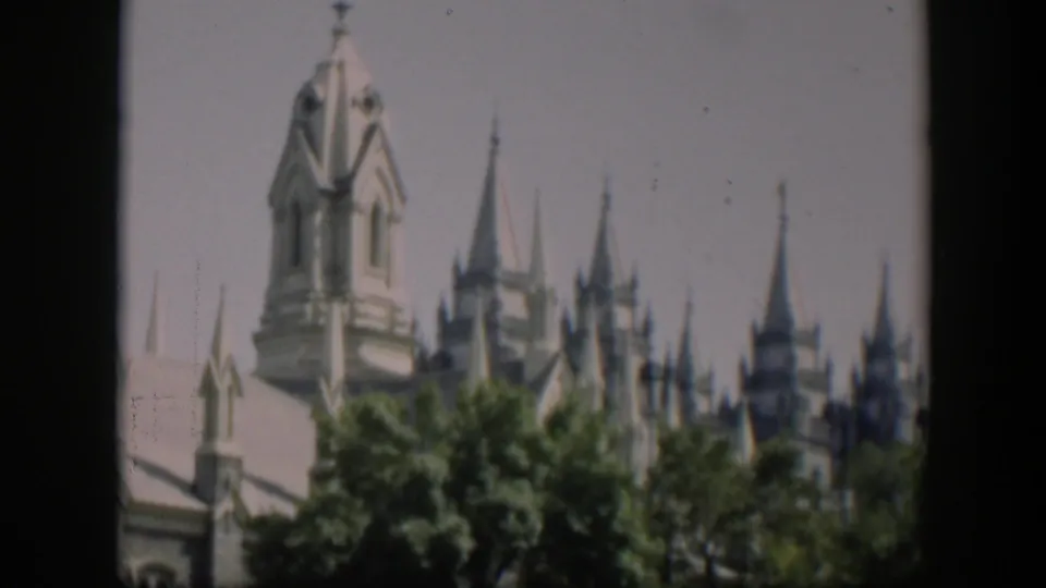 A pale, ornate building with multiple spires appears beyond green trees in a 1951 Wyoming home-movie frame.