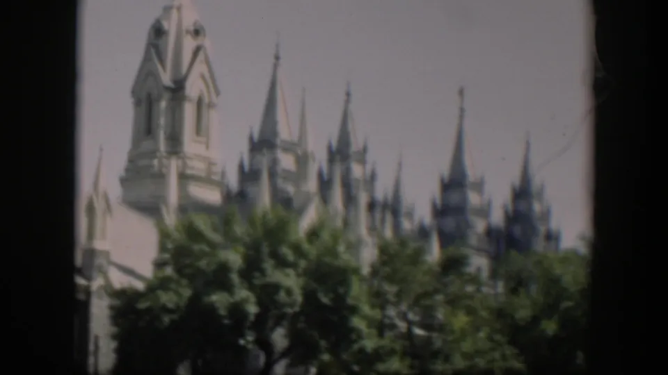 1951 8mm view of a tall white tower and clustered spires partly obscured by mature trees.