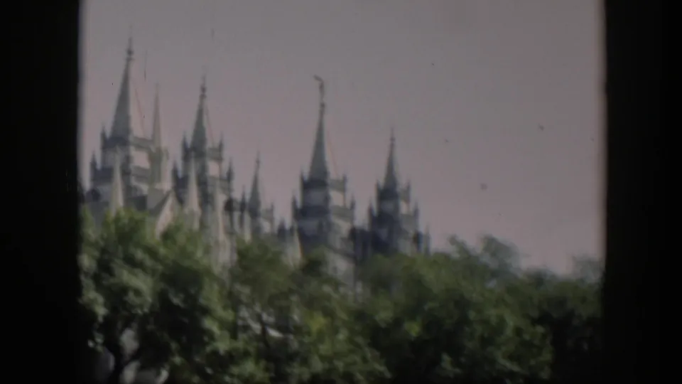 Distant 8mm view of a multi-spired religious landmark partly hidden by trees, with a dark vertical film border at left.
