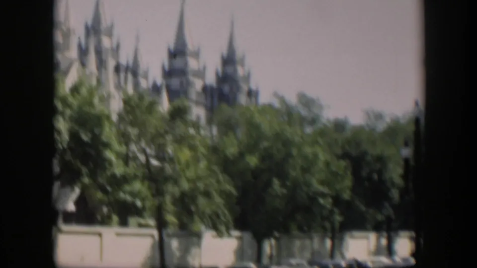 1951 home-movie frame showing spires above dense trees, with a low wall and a parked car visible near the bottom edge.