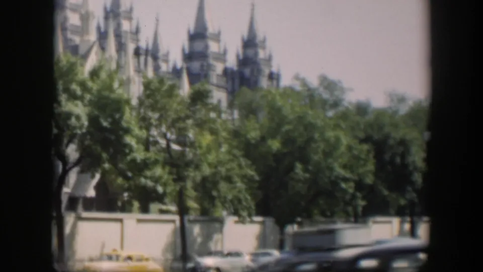 Soft 8mm view of a spired religious landmark beyond trees, with a pale wall and parked cars along the foreground street.