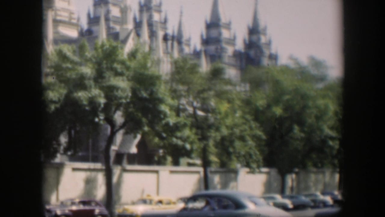 Cars pass a tan Gothic-style building partly hidden by trees.
