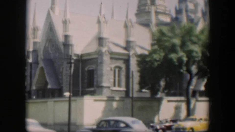 1951 Wyoming street frame showing cars below a tan building with a pointed entrance, spires, trees, and bright sun.
