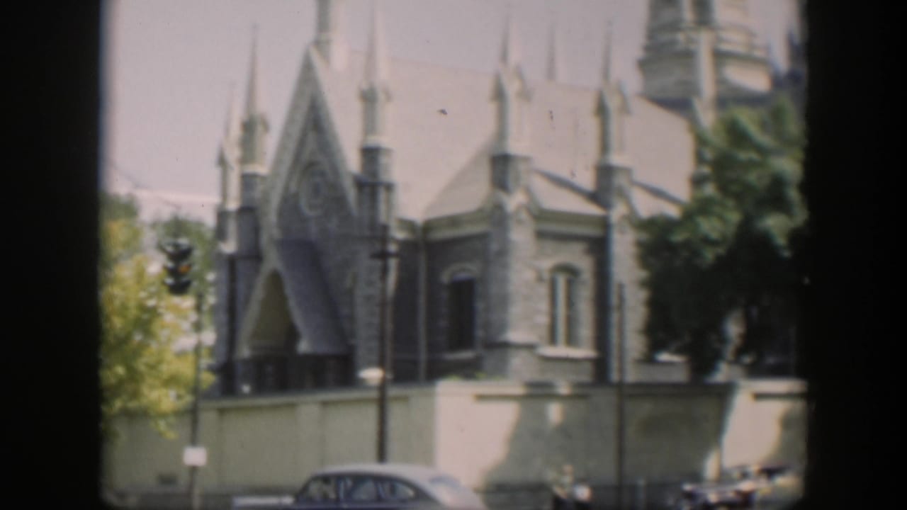1951 8mm home-movie frame of a stone Gothic-style church exterior in Wyoming, with spires, a low wall, trees, and cars along the street.