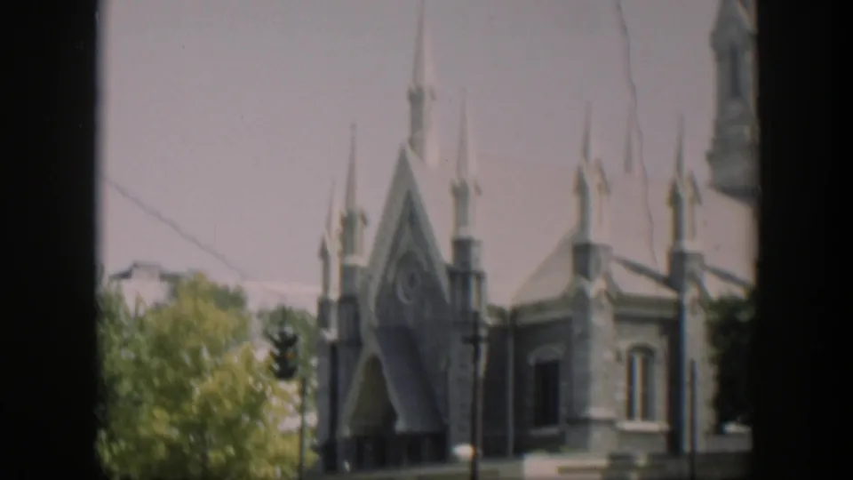 1951 8mm frame of a Gothic-style church entrance and side wall, with pointed arches, narrow spires, trees, and street lighting visible.