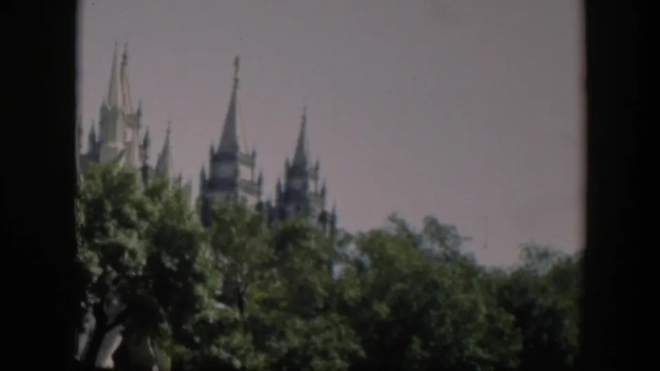 Soft 8mm frame showing Gothic-style spires above a line of leafy trees, with the church partly hidden from the street view.