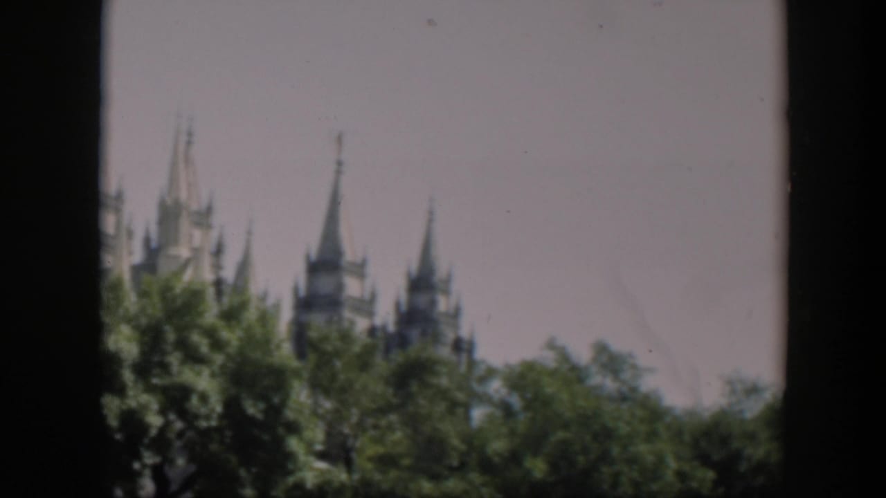 1951 Wyoming 8mm frame showing distant religious spires and roof peaks above leafy trees under a pale sky.