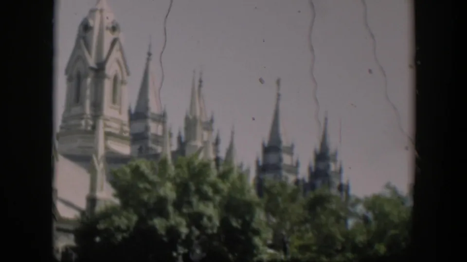 1951 Wyoming home-movie frame with a pale religious tower and multiple pointed spires partly obscured by trees.