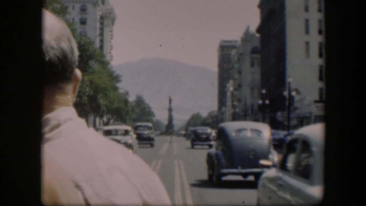 City street seen from behind a seated person, with older cars and buildings ahead.