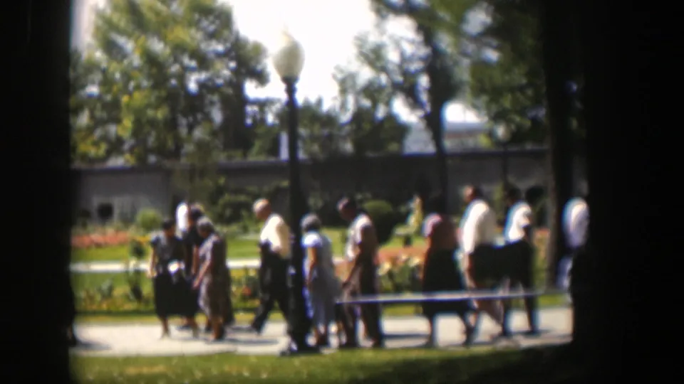 Wyoming 1951 park frame showing pedestrians on a walkway near globe lampposts, trees, lawn, and low flower beds.
