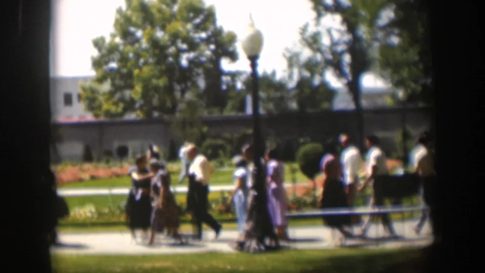 1951 Wyoming 8mm frame of adults in mid-century clothing walking past lawns, orange flowerbeds, trees, and a lamppost.