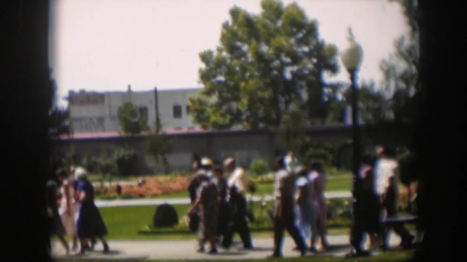 Adults in 1951 Wyoming park footage walk past landscaped lawns and orange flowerbeds with trees and a low building behind them.