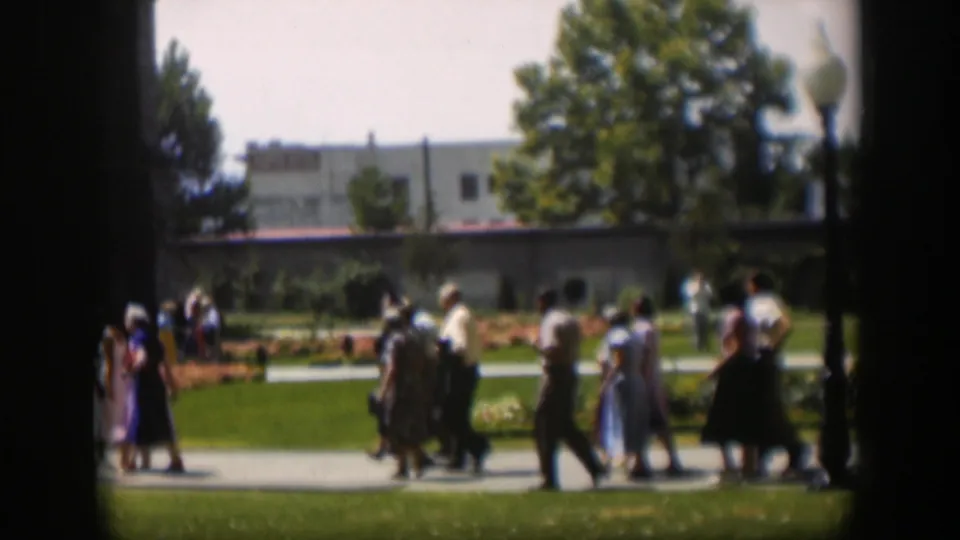 Mid-century pedestrians in Wyoming walk through a park scene with orange flowerbeds, grass, trees, and a building in the background.