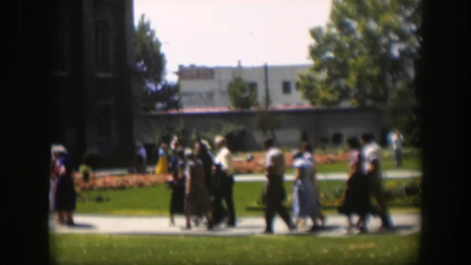 1951 Wyoming home-movie frame showing adults walking beside orange flowerbeds and lawns, with trees, a lamppost, and building beyond.