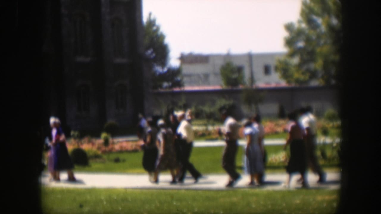 1951 Wyoming home-movie frame showing adults in dresses and shirts walking outdoors along a path near lawn and flowerbeds.