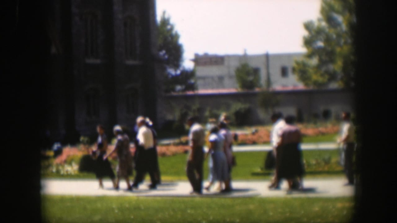 Several adults in mid-century clothing walk in small groups across a sunny lawn-side path.